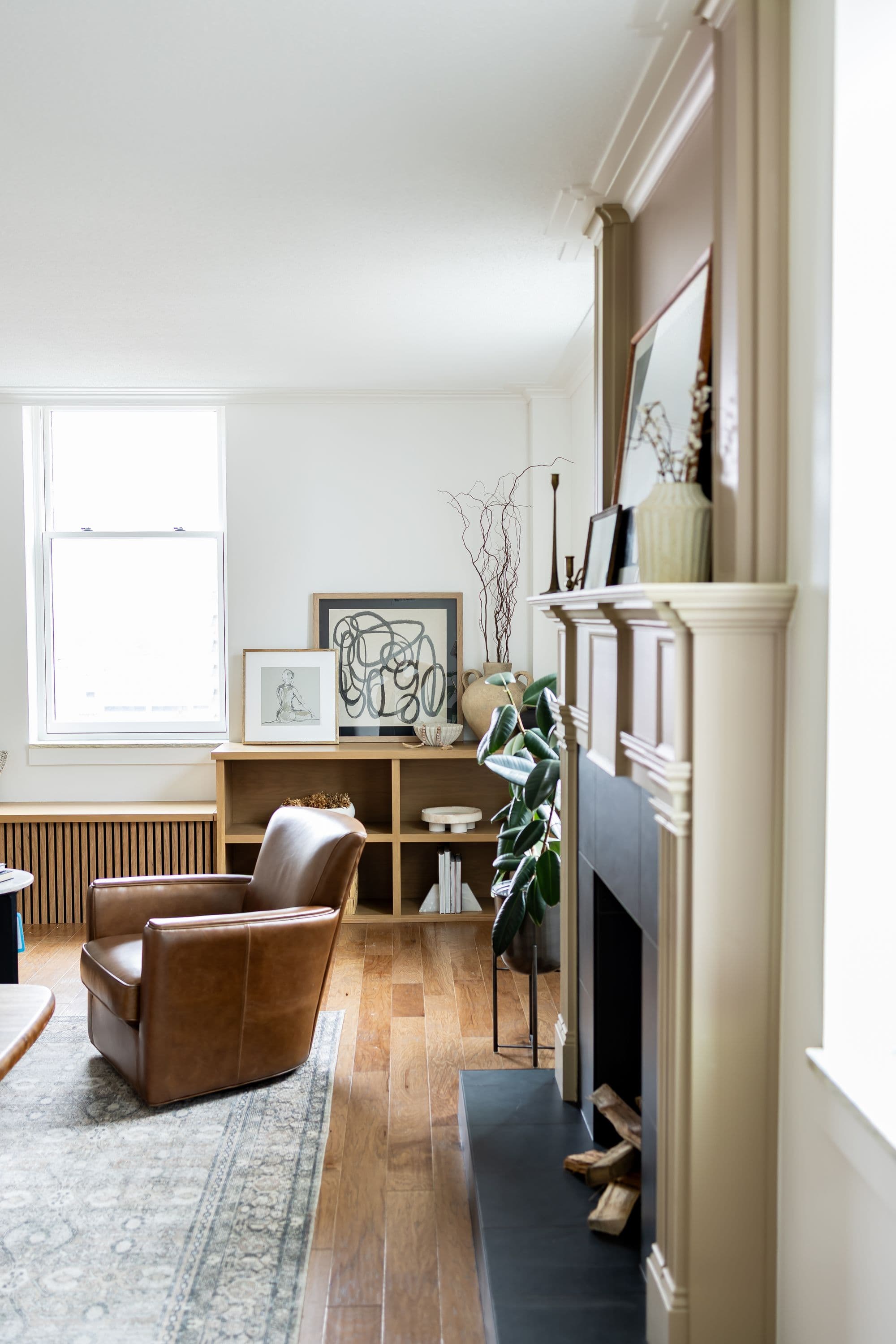 A stylish living room showcases a brown leather armchair, a bookshelf with decorative items, and a fireplace with a plant beside it. The room features hardwood floors and natural light from a window.