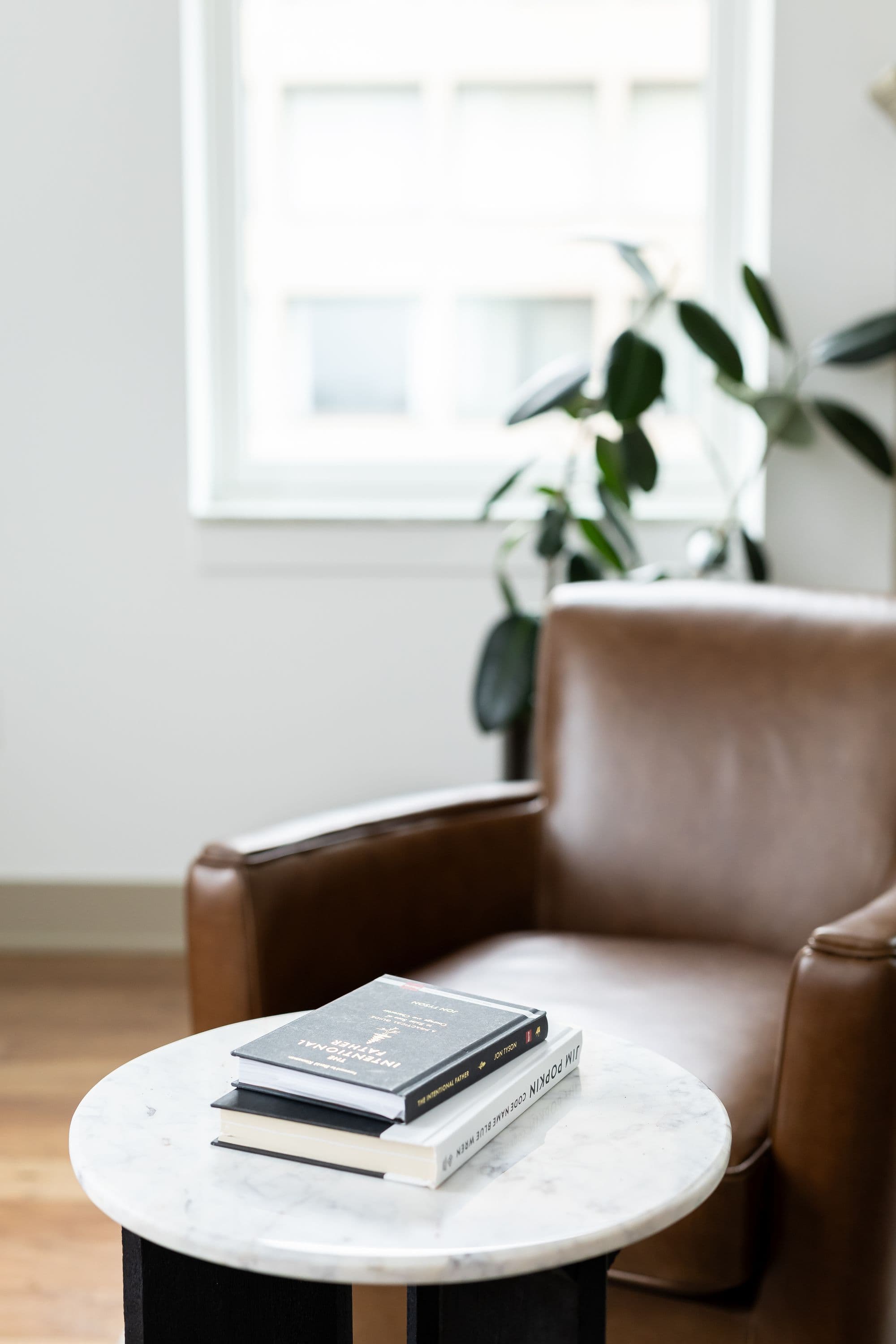 Interior shot of a living room featuring a brown leather armchair, marble-topped side table with two books, and a green leafy plant. The room has light wooden floors and natural light from a window.