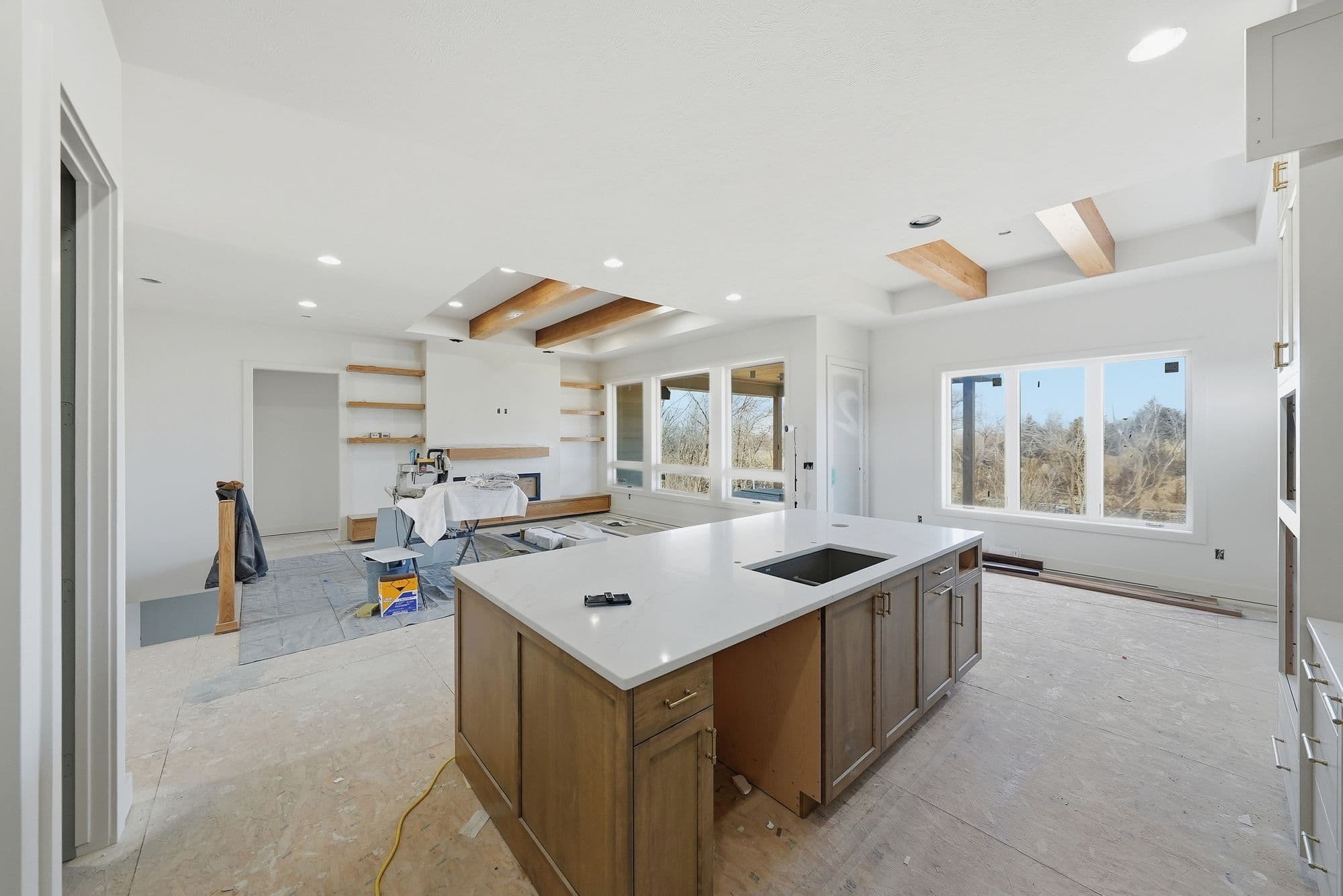 Unfinished kitchen area featuring a large center island with a white countertop and sink, along with unfinished wood cabinets. There are large windows looking out to a wooded area, and exposed wood beams on the ceiling.