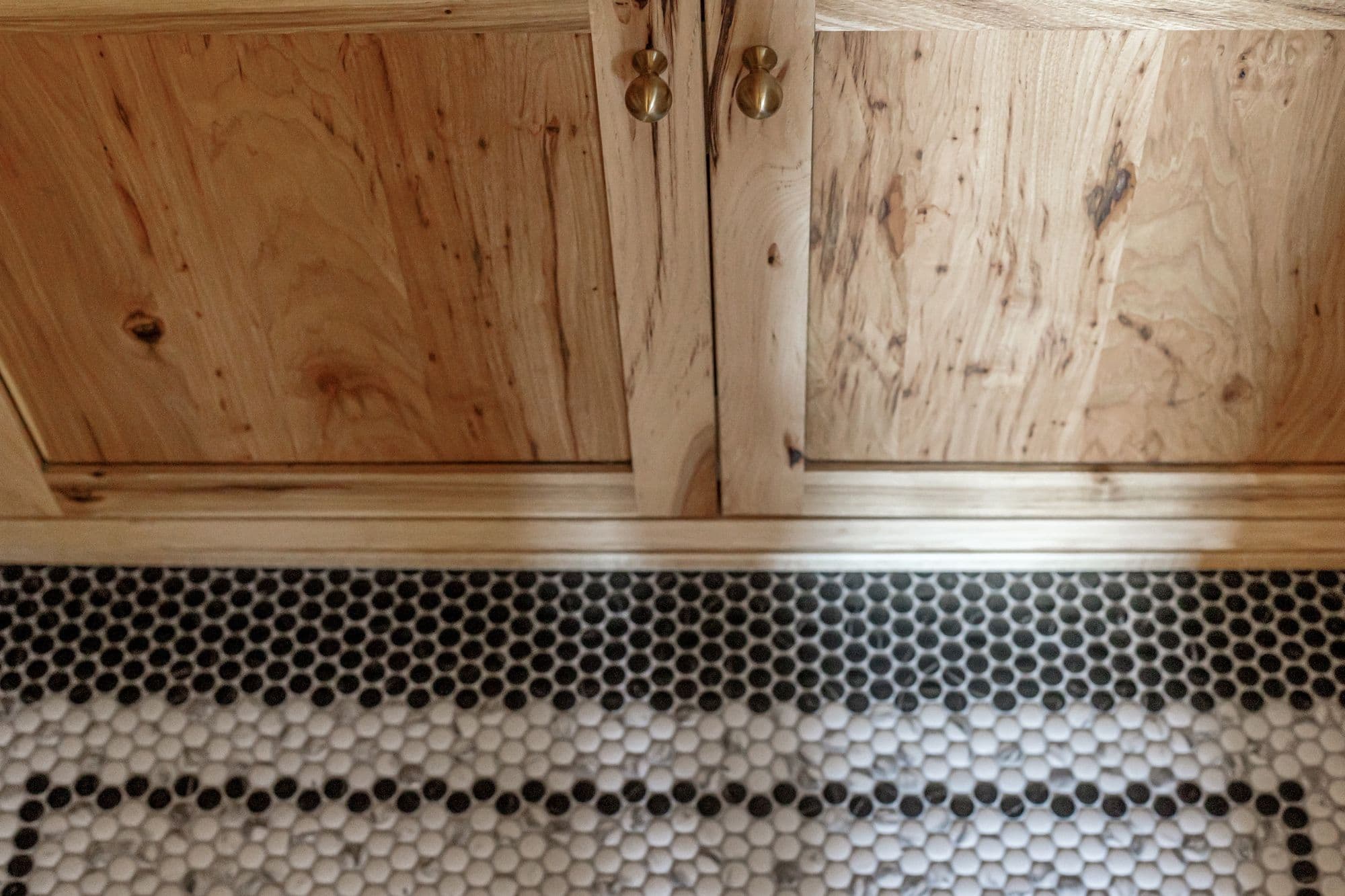 Bathroom vanity with natural wood cabinets and brass hardware sits above a black and white penny tile floor. The penny tile floor features a black border and white with black speckled tiles.