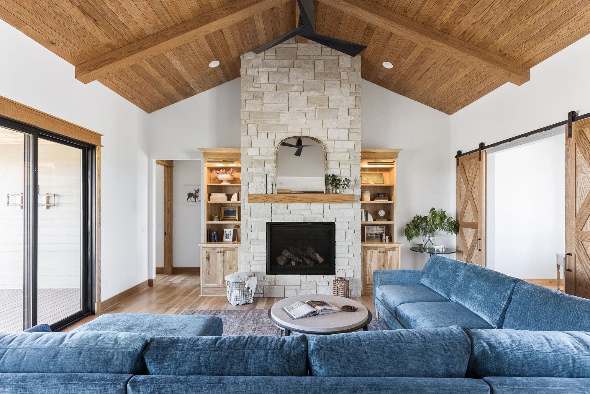 Bright living room with vaulted wood ceiling, stone fireplace with built-in shelves, and a large blue sectional sofa. Sliding barn doors and a modern sliding glass door offer access to other areas.