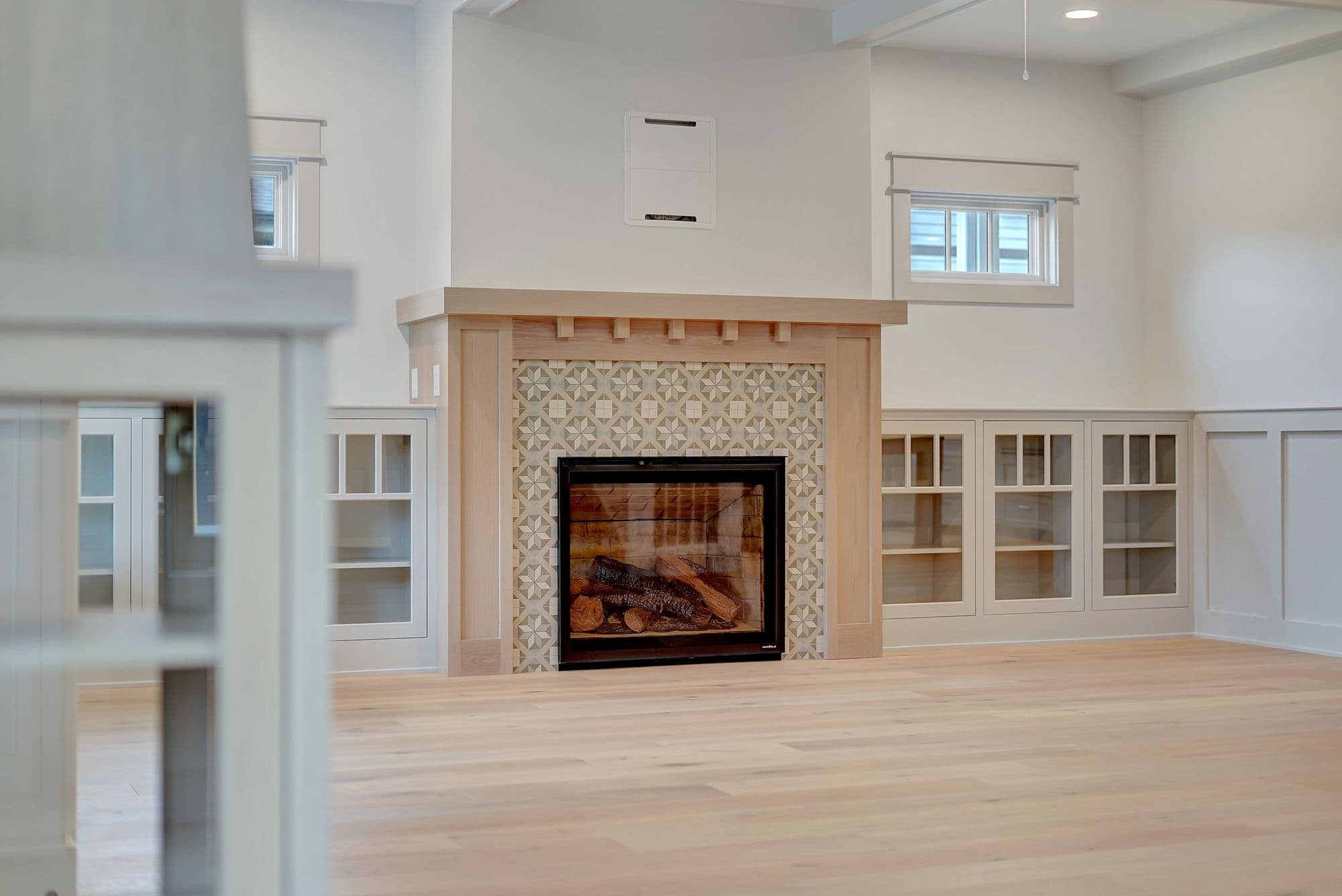 Bright living room features hardwood floors, built-in cabinets with glass doors, and a fireplace with decorative tile surround. A window with soft light is visible on the right wall.